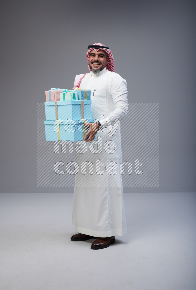 Saudi man Wearing Thob and red Shomag standing holding gift box on Gray background
