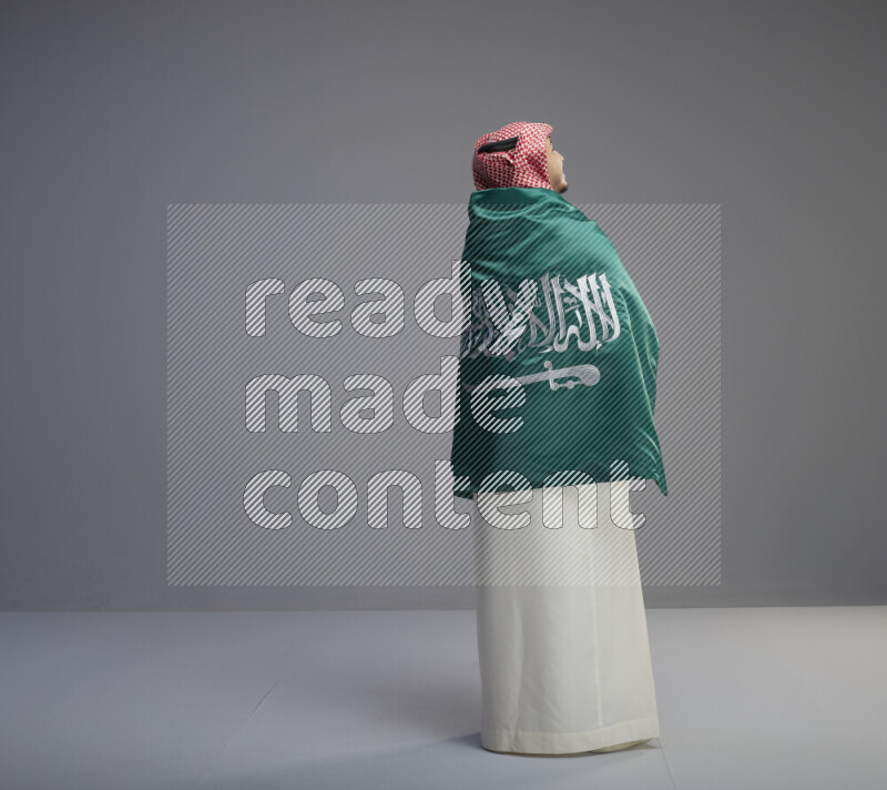 A Saudi man standing wearing thob and red shomag wrapping big saudi flag on gray background