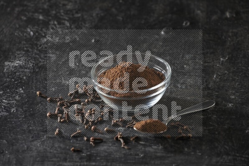 A glass bowl and a metal spoon full of cloves powder with gloves grains beside them on a textured black flooring