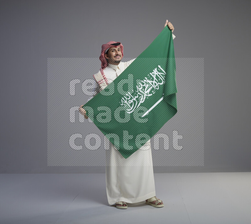 A saudi man standing wearing thob and red shomag holding big saudi flag on gray background