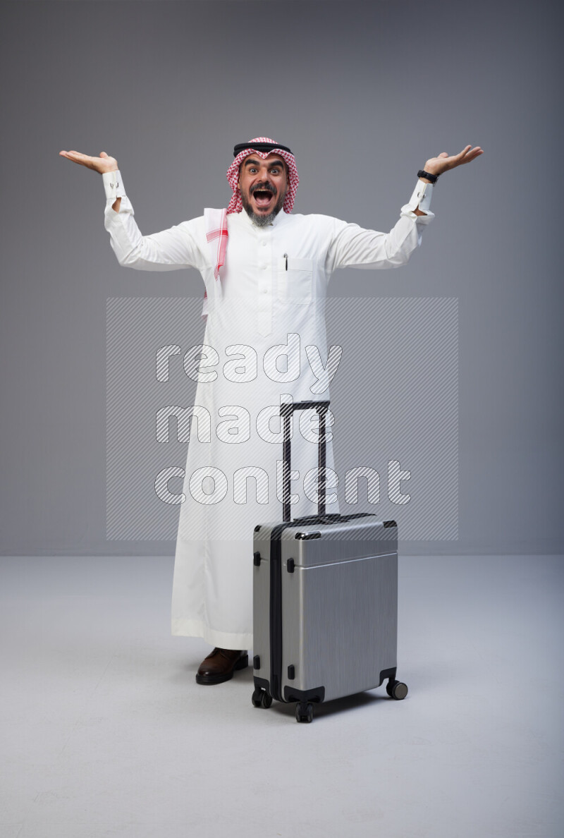 Saudi man wearing Thob and red Shomag standing holding Travel bag on Gray background