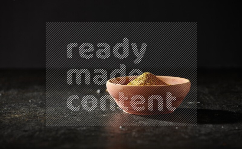 A wooden bowl full of turmeric powder with dried turmeric fingers on textured black flooring