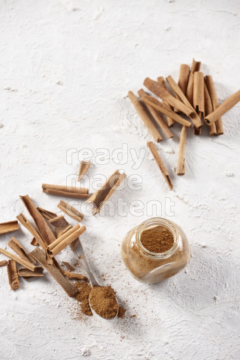 Herbal glass jar full cinnamon powder and a metal spoon surrounded by cinnamon sticks on a white background