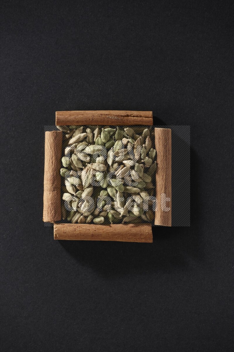 أربعة من أعواد القرفة مكونة شكل مربع بداخلها حبوب الهيل علي خلفية سوداء