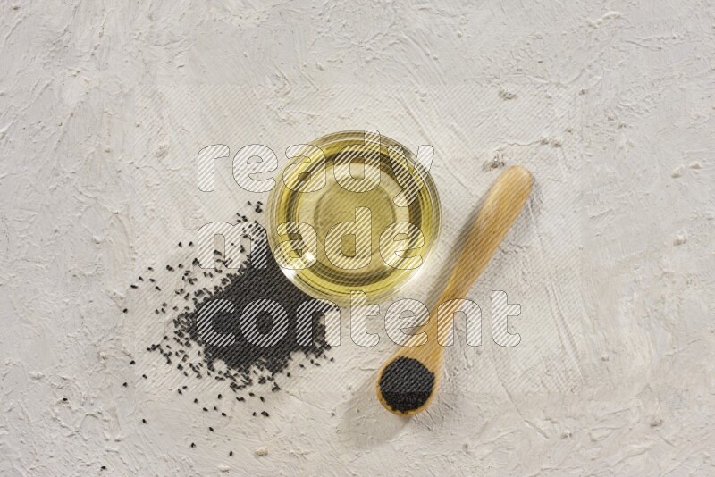 A glass bowl full of black seeds oil and wooden spoon full of black seeds with seeds spread on a textured white flooring