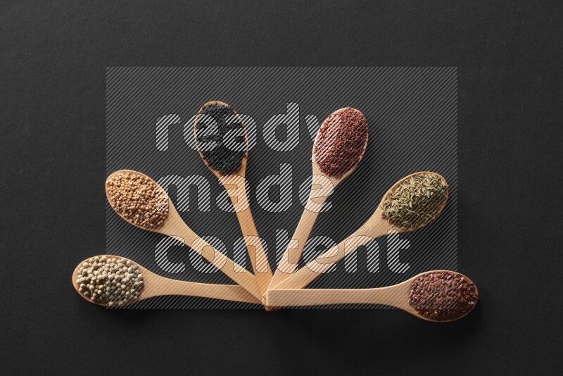 wooden spoons filled with white peppers, mustard seeds, black seeds, garden cress, cumin and flax on black flooring