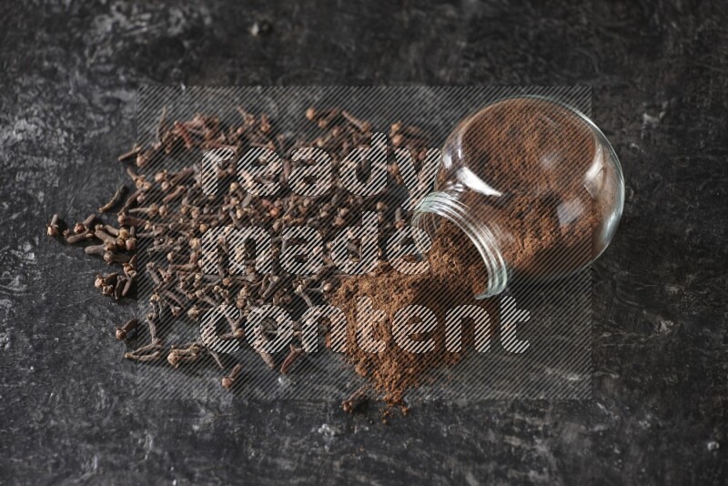 A flipped glass spice jar full of cloves powder and powder came out of it with cloves spread on textured black flooring