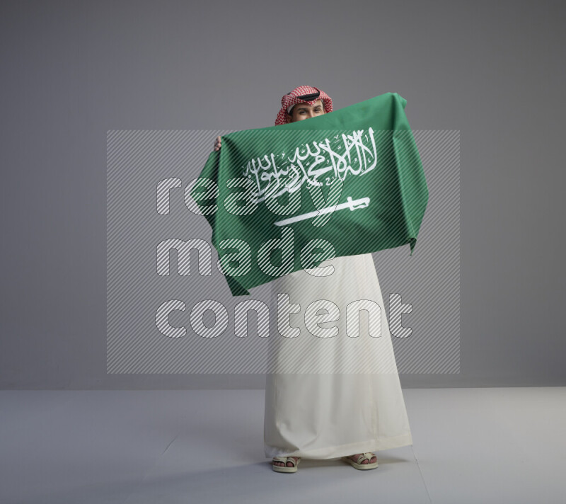 A saudi man standing wearing thob and red shomag holding big saudi flag on gray background