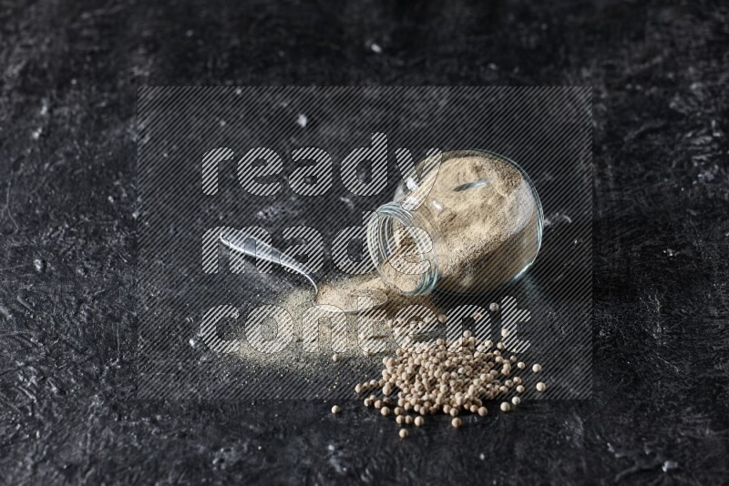 A flipped herbal glass jar and metal spoon full of white pepper powder with spilled powder and pepper beads on textured black flooring