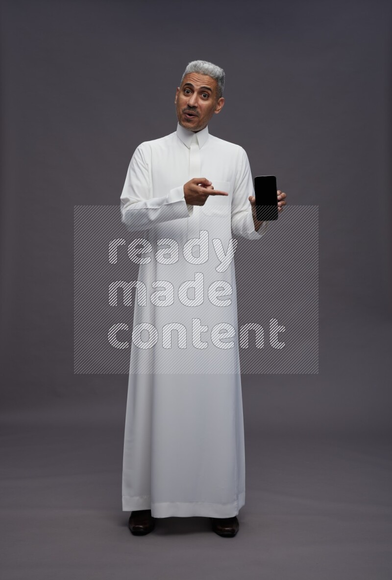 Saudi man wearing thob standing showing phone to camera on gray background
