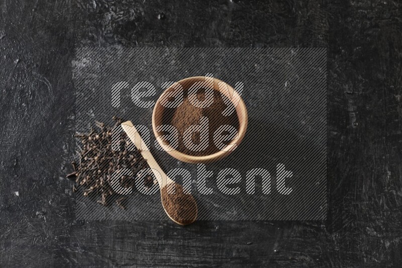 A wooden bowl and a wooden spoon full of cloves powder with spreaded cloves on a textured black flooring