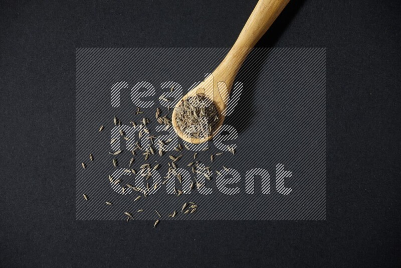 A wooden spoon full of cumin seeds on black flooring