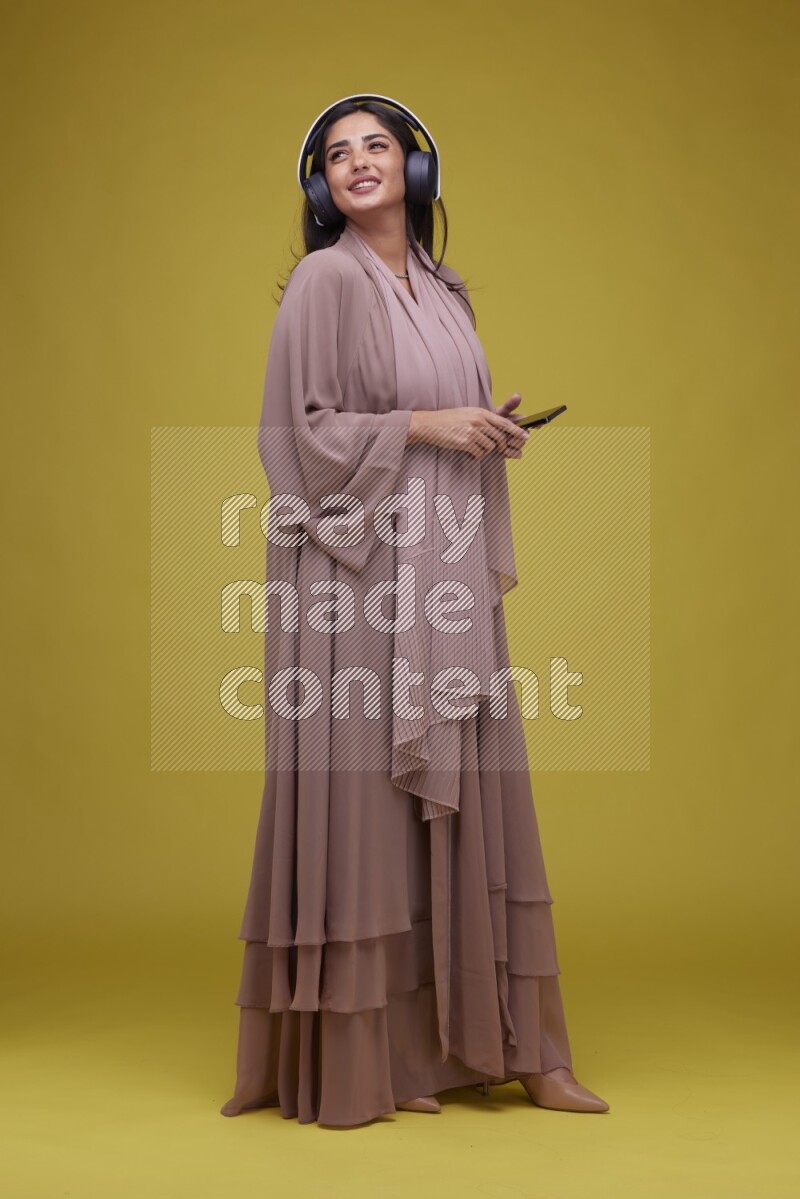 A Saudi woman Listing to Music on a Yellow Background wearing Brown Abaya