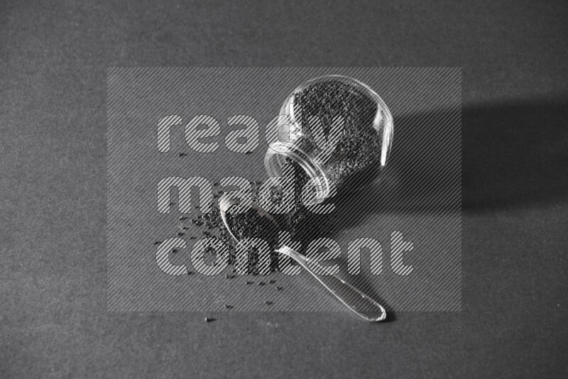 A flipped glass spice jar full of black seeds and the seeds spread out with a metal spoon full it on a black flooring