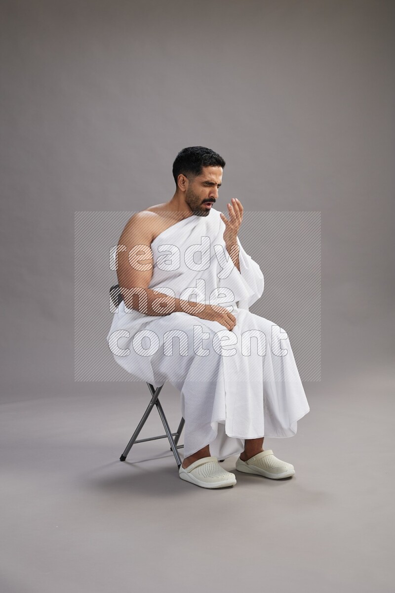 A man wearing Ehram sitting on chair showing how to sneeze on gray background