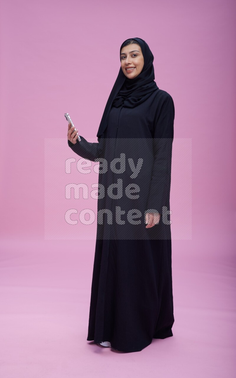 Saudi woman wearing Abaya standing taking selfie on pink background