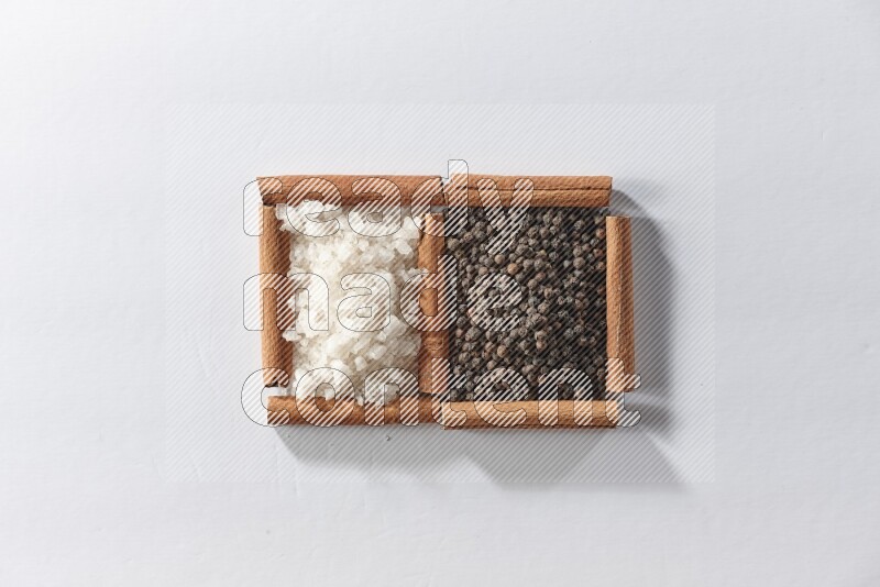 مربعان من أعواد القرفة ممتلئان بحبوب الفلفل الأسود والملح الأبيض علي خلفية بيضاء