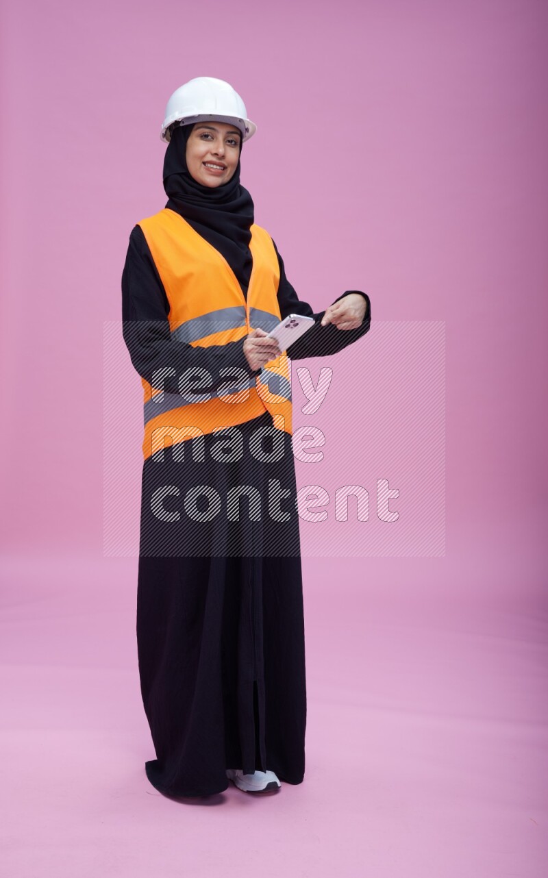 Saudi woman wearing Abaya with engineer vest and helmet standing texting on phone on pink background