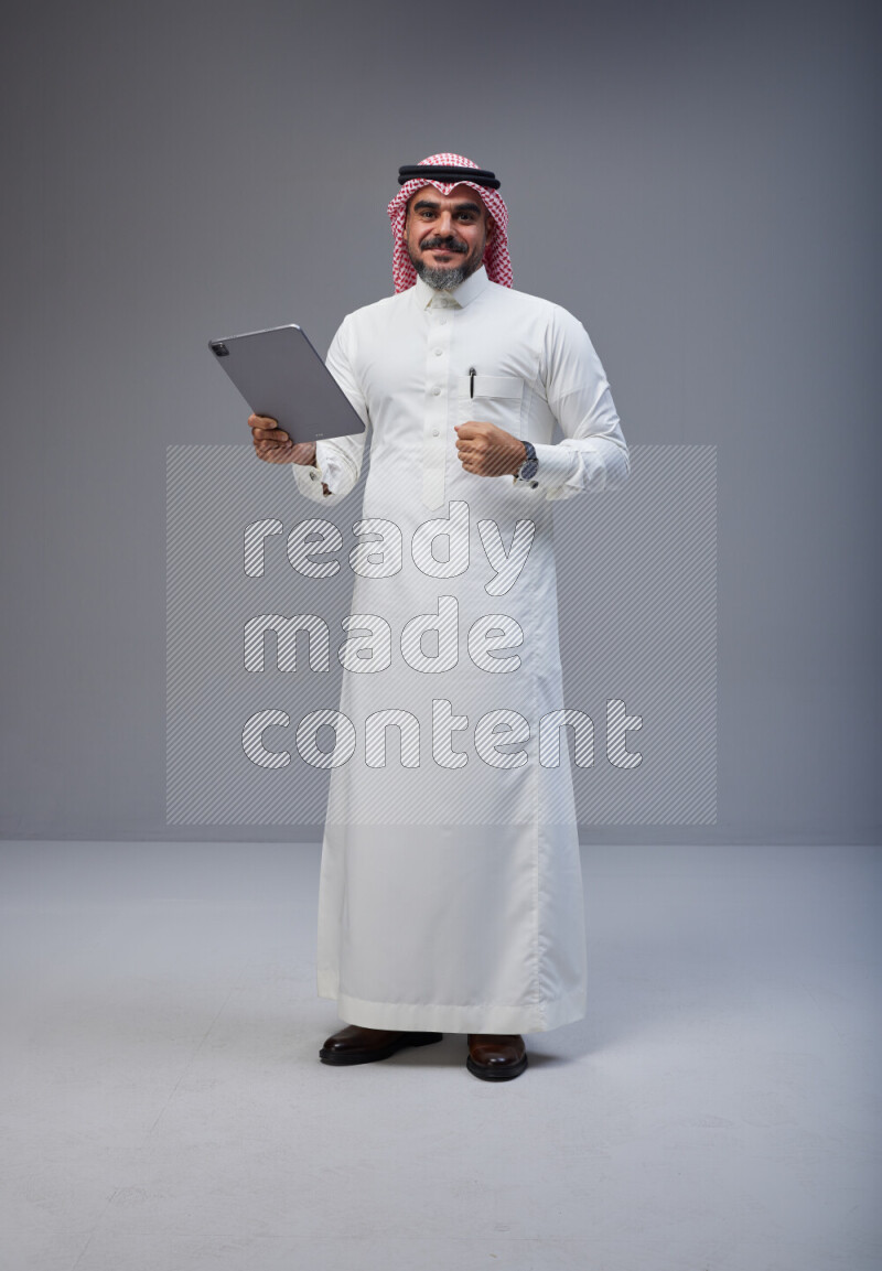 Saudi man Wearing Thob and red Shomag standing working on tablet on Gray background