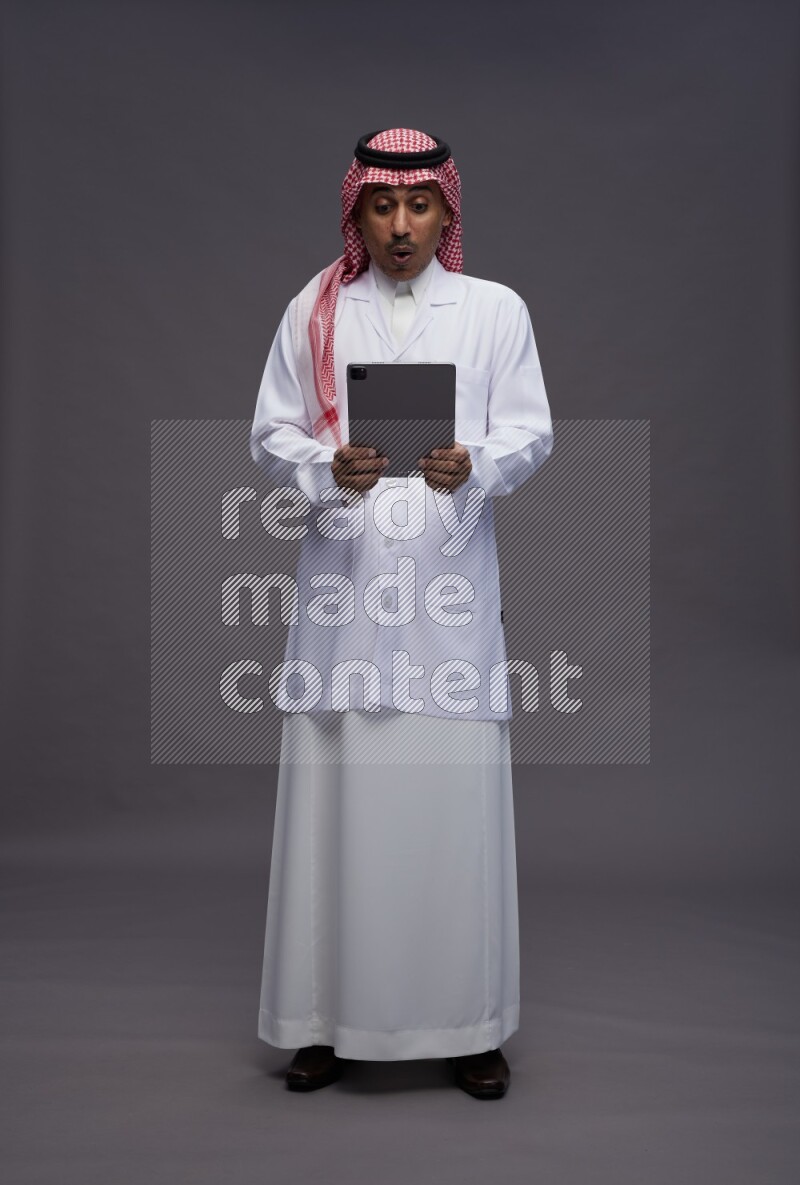 Saudi man wearing thob with lab coat and shomag with pocket employee badge standing working on tablet on gray background