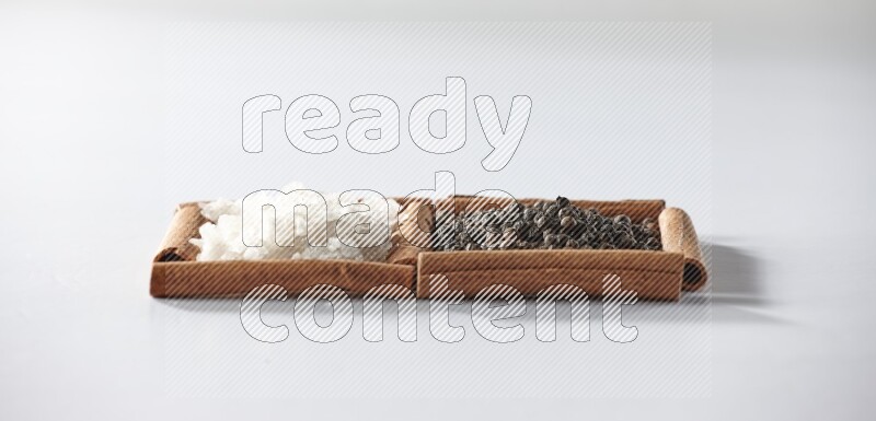 مربعان من أعواد القرفة ممتلئان بحبوب الفلفل الأسود والملح الأبيض علي خلفية بيضاء