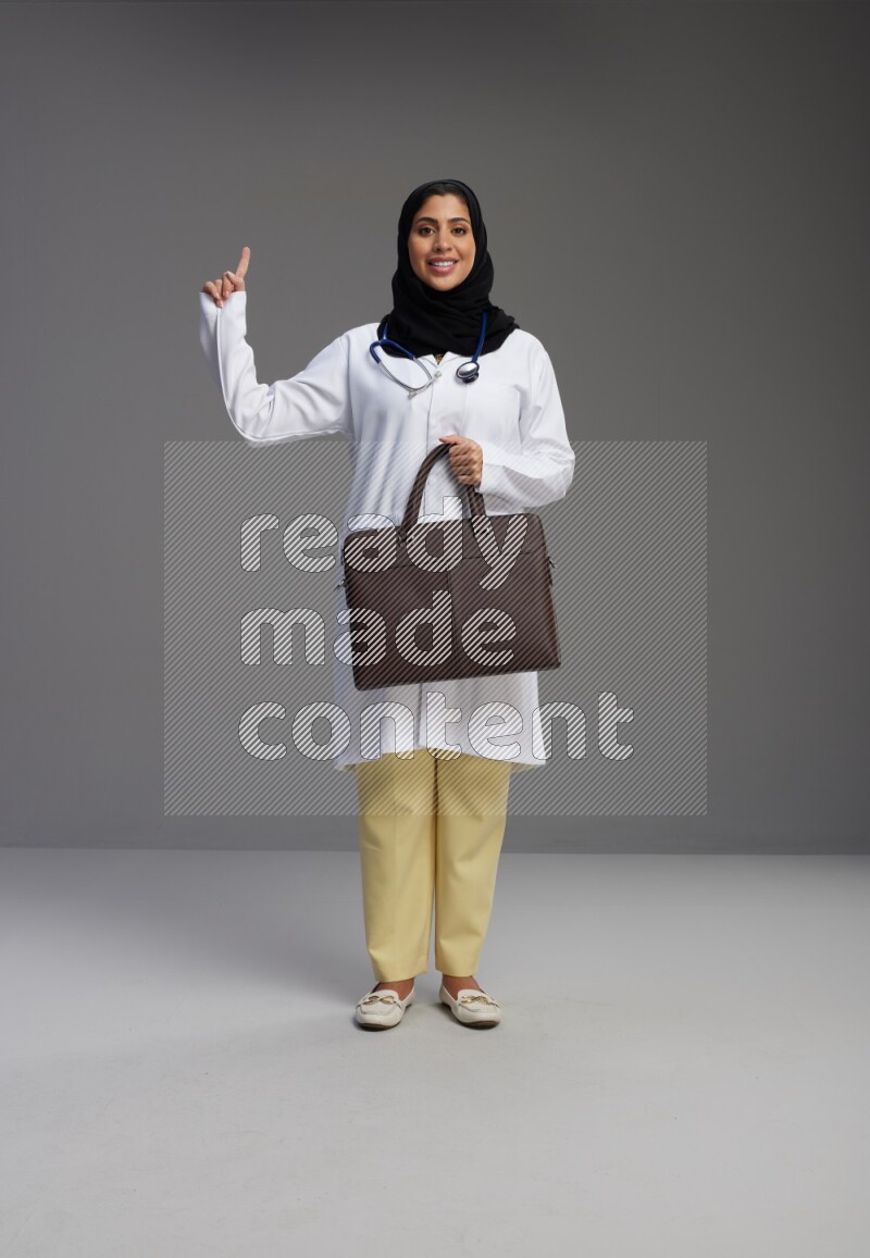 Saudi woman wearing lab coat with stethoscope standing holding bag on Gray background