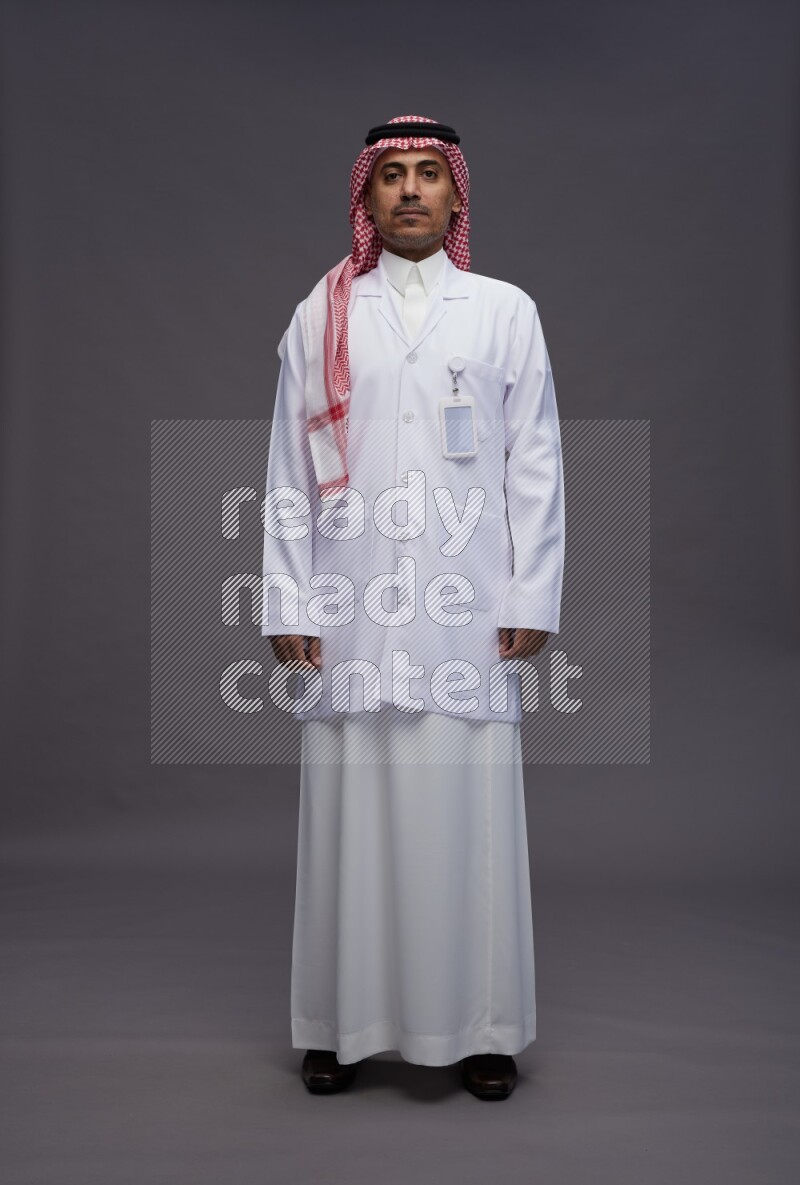 Saudi man wearing thob with lab coat and shomag with pocket employee badge standing interacting with the camera on gray background