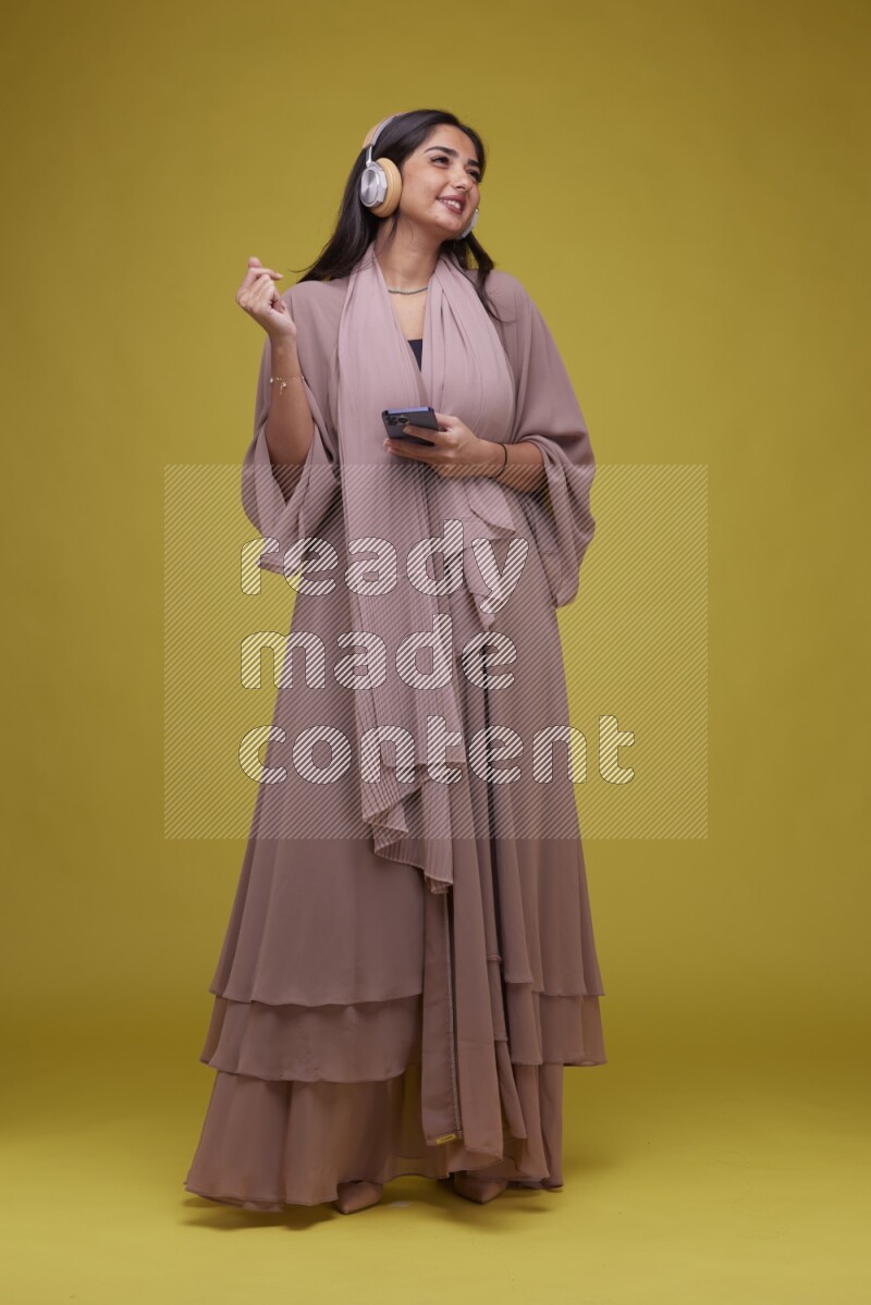 A Saudi woman Listing to Music on a Yellow Background wearing Brown Abaya