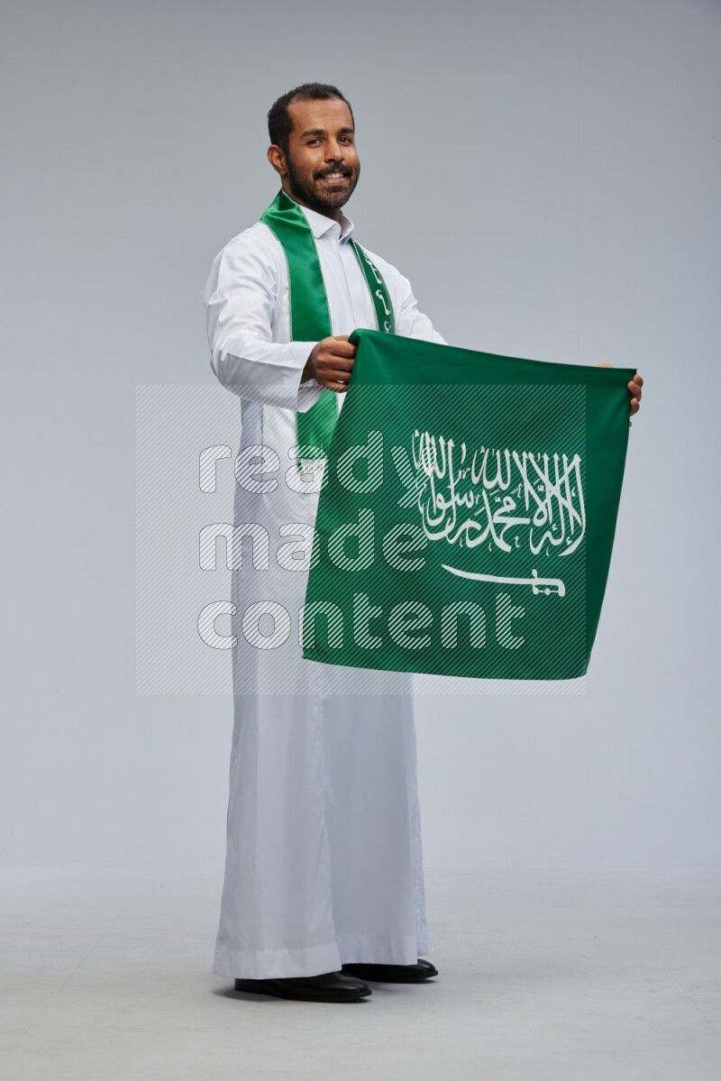 Saudi man wearing Thob standing holding Saudi flag on Gray background