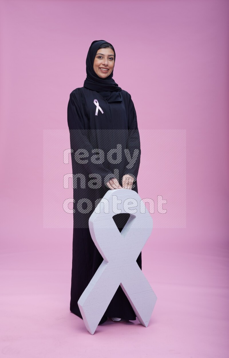 Saudi woman wearing pink ribbon on abaya standing holding awareness ribbon on plain pink background