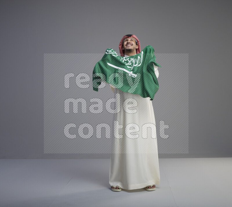 A saudi man standing wearing thob and red shomag holding big saudi flag on gray background