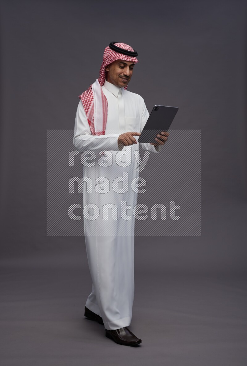 Saudi man wearing thob and shomag standing working on tablet on gray background