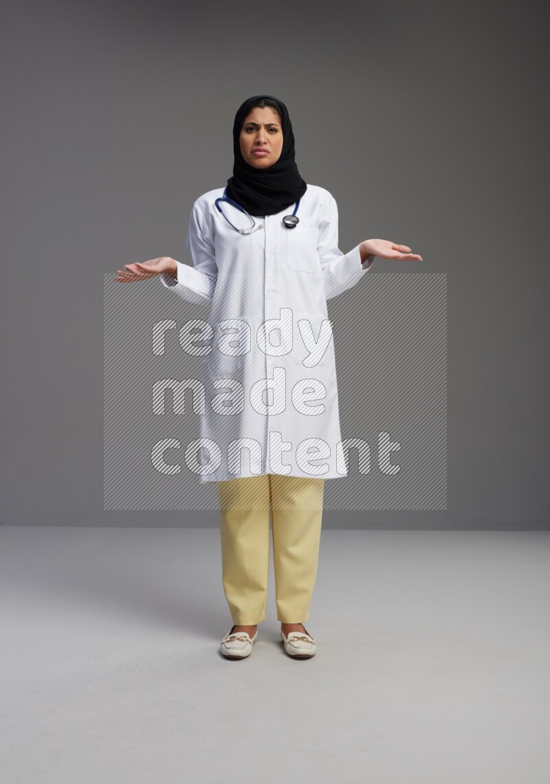 Saudi woman wearing lab coat with stethoscope standing interacting with the camera on Gray background