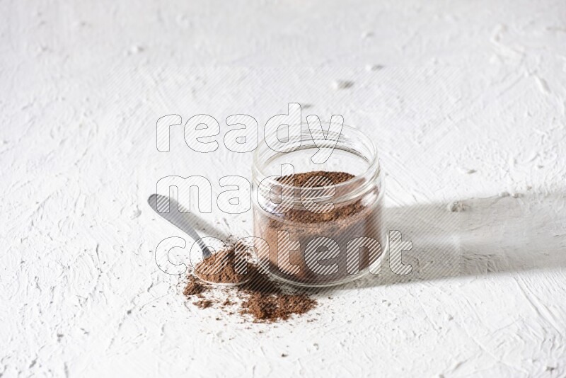 A glass jar full of cloves powder with a metal spoon on a textured white flooring