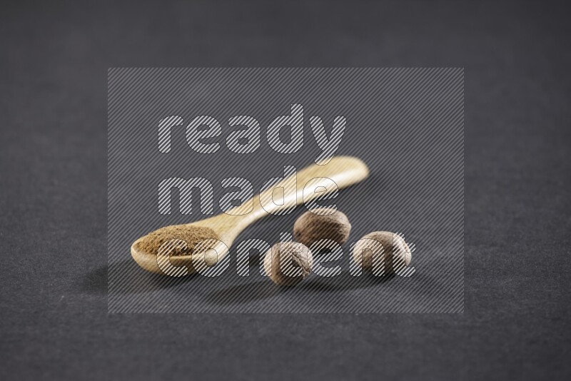 ملعقة خشبية ممتلئة ببودرة جوزة الطيب مع حبوب جوزة الطيب الكاملة بجانبها على خلفية سوداء