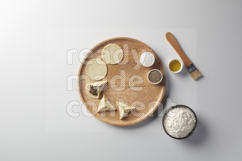 two closed sambosas and one open sambosa filled with meat while flour, salt, black pepper and oil with oil brush aside in a wooden dish on a white background