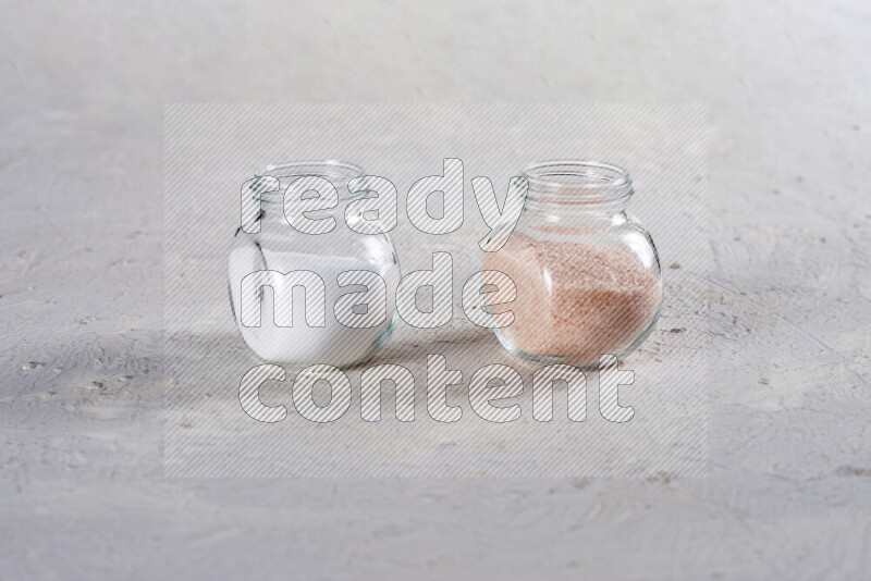 2 glass jars one is full of fine himalayan salt and the other with table salt on white background
