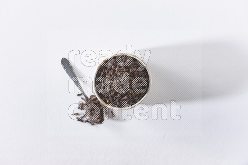 A beige ceramic bowl full of cloves and a metal spoon next to it on a white flooring