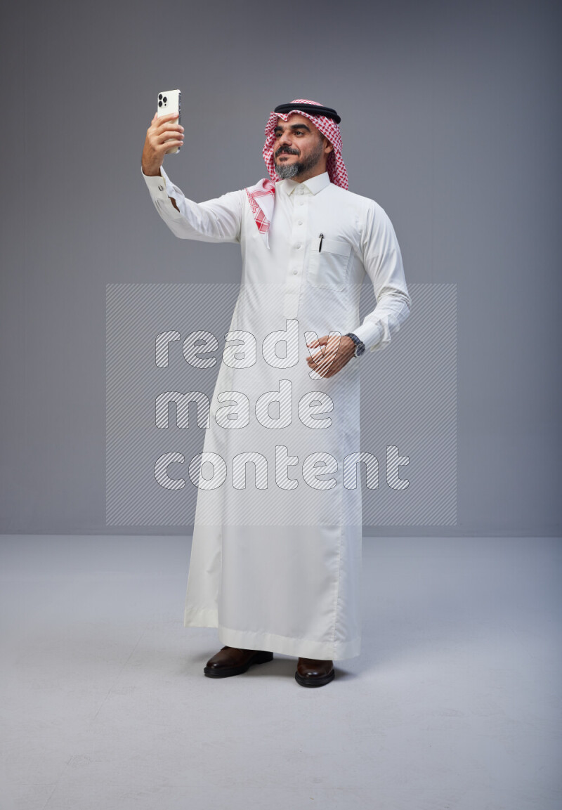 Saudi man Wearing Thob and red Shomag standing taking selfie on Gray background