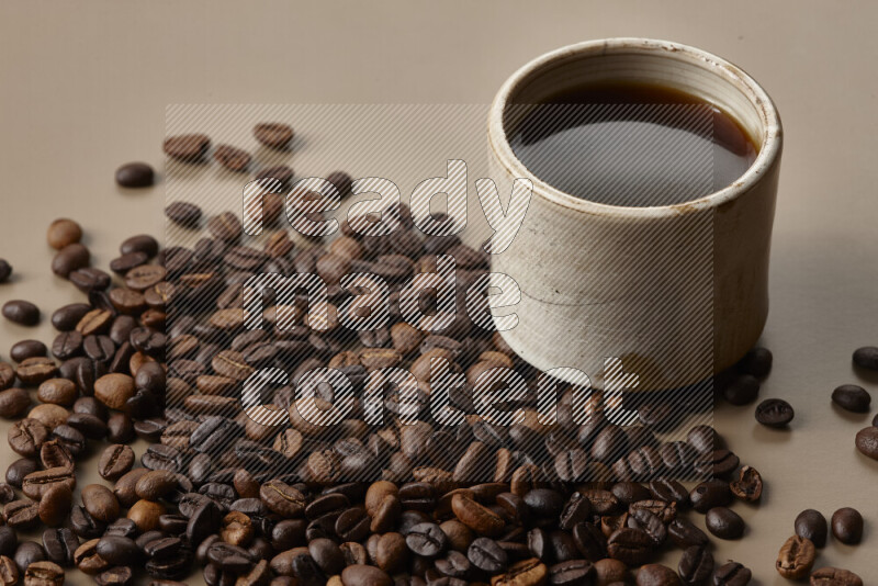 A beige pottery cup of coffee surrounded by roasted coffee beans on beige background