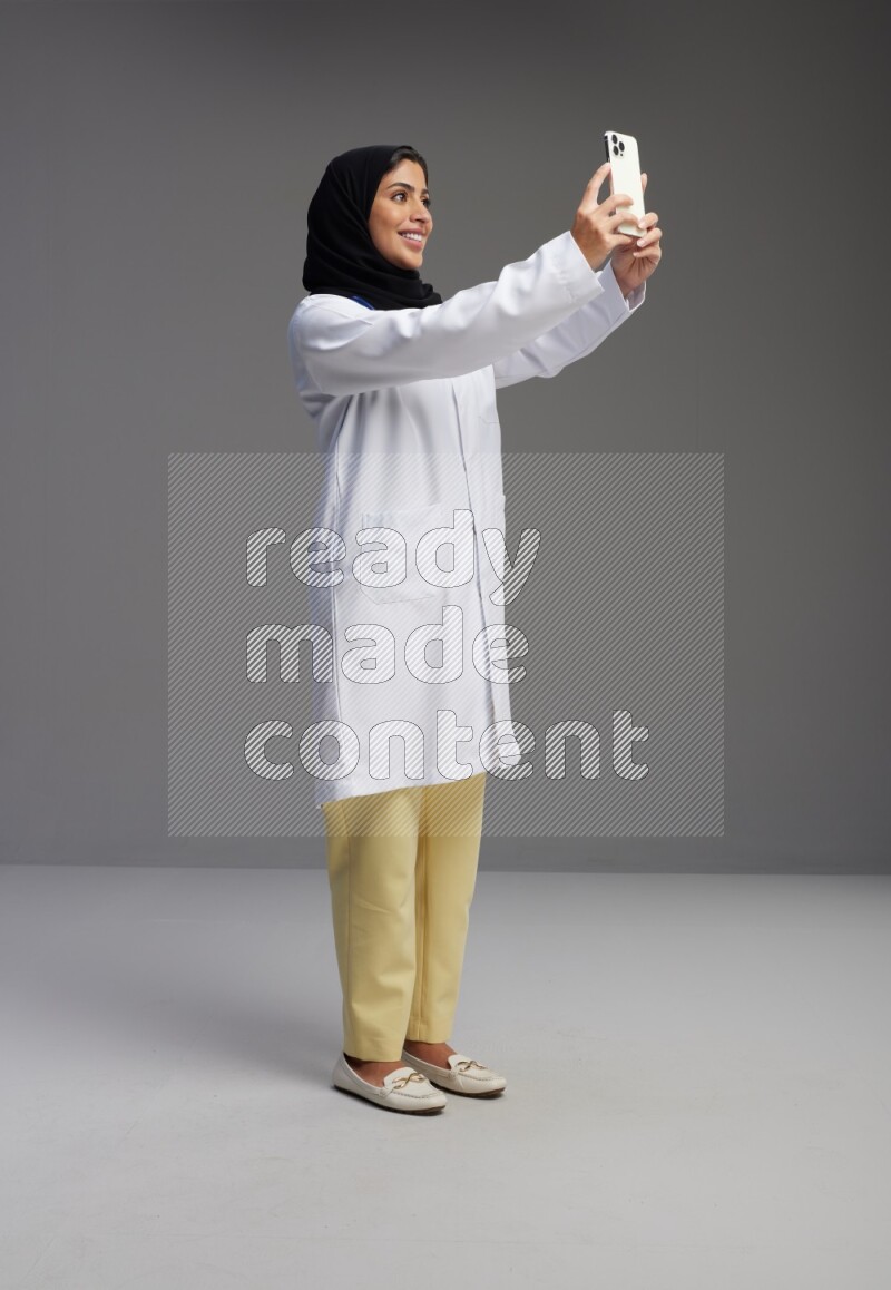 Saudi woman wearing lab coat with stethoscope standing taking selfie on Gray background