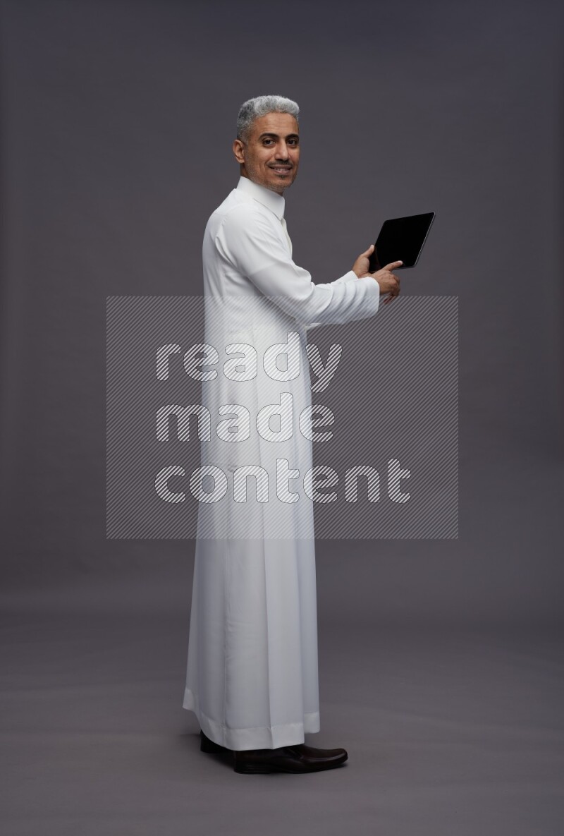 Saudi man wearing thob standing working on tablet on gray background