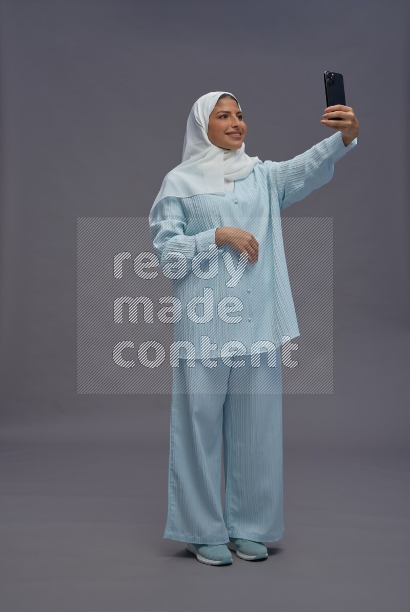 Saudi woman wearing hijab clothes standing taking selfie on gray background