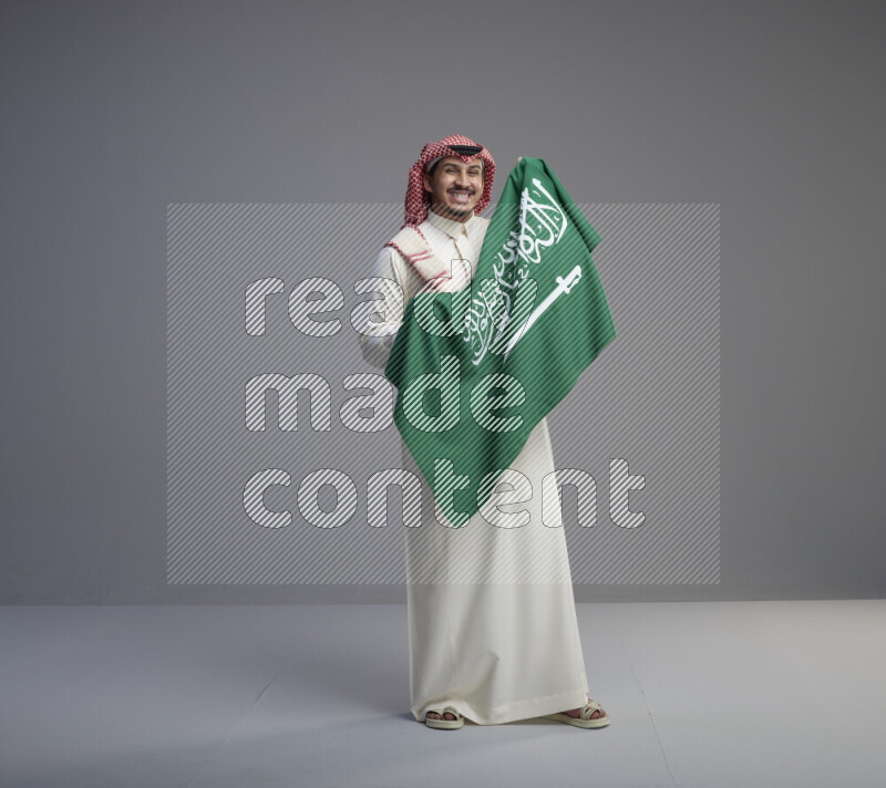 A saudi man standing wearing thob and red shomag holding big saudi flag on gray background