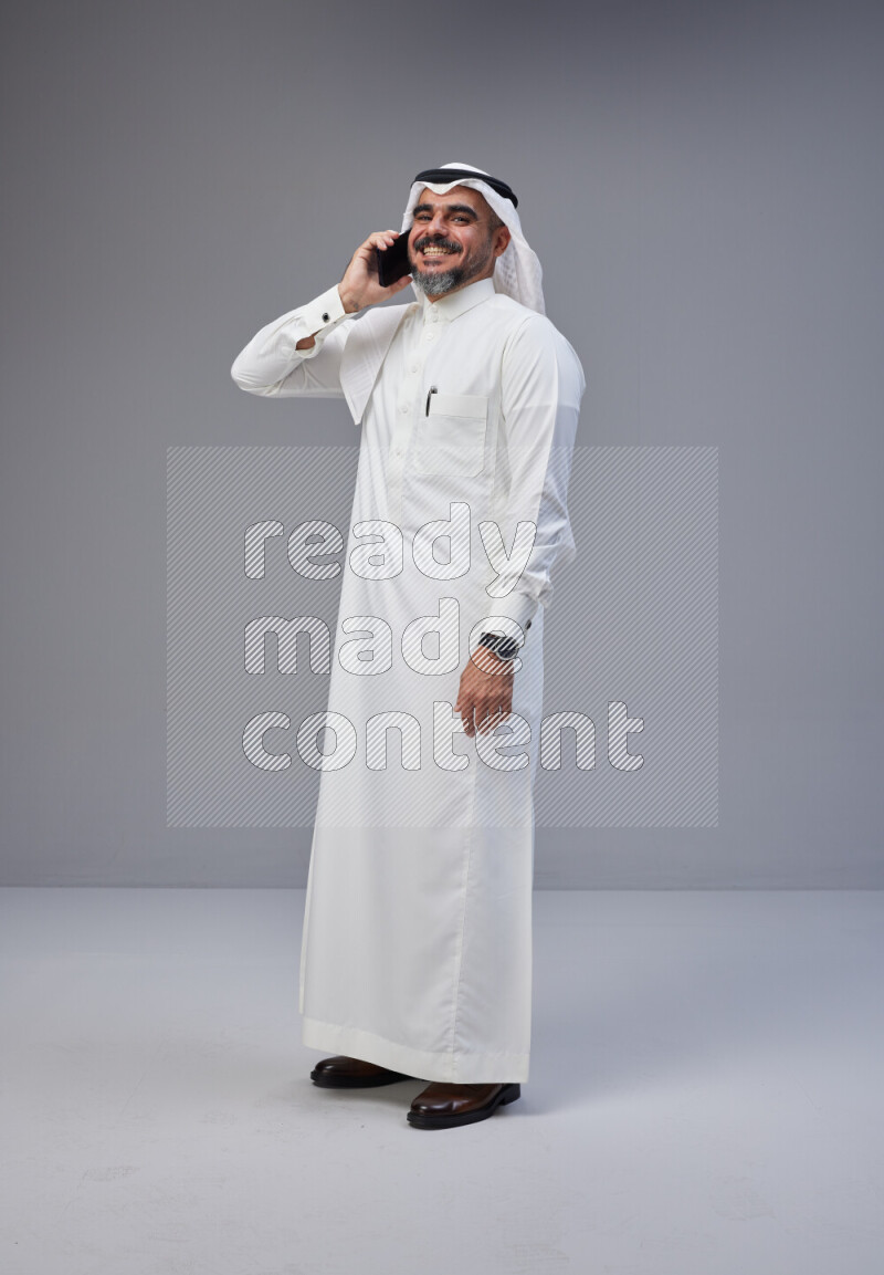 Saudi man Wearing Thob and white Shomag standing talking on phone on Gray background