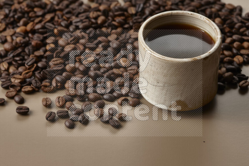 A beige pottery cup of coffee surrounded by roasted coffee beans on beige background