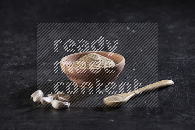 A wooden bowl and spoon full of garlic powder and beside it garlic cloves on a textured black flooring