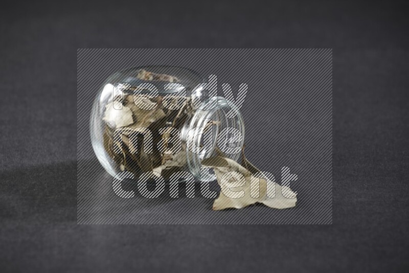 A glass spice jar full of dried bay leaves and jar is flipped and leaves spread out on black flooring