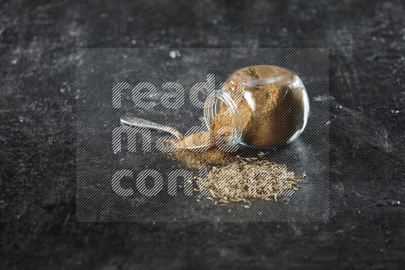 A flipped glass spice jar and a metal spoon full of cumin powder and powder spilled out with cumin seeds on a textured black flooring