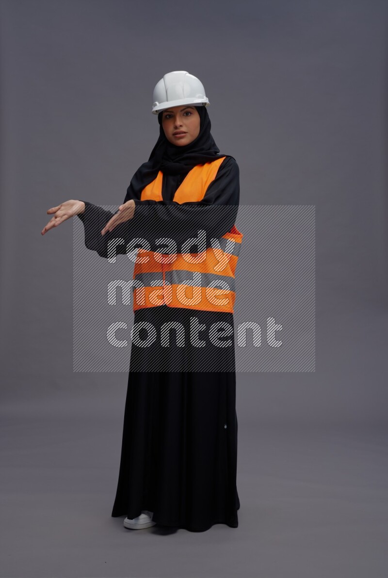 Saudi woman wearing Abaya with engineer vest standing interacting with the camera on gray background